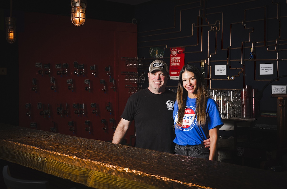 A man and a woman stand together behind a textured, industrial-style bar in a dimly lit space. The man, wearing a black t-shirt and a baseball cap, has his arm around the woman, who is wearing a bright blue "Greek's Pizzeria" t-shirt. The background features a dark wall decorated with a network of copper pipes, small framed quotes, and rows of what look like tap handles or keys. A warm, vintage-style industrial light fixture hangs above them.