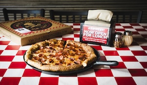 A fresh, thick-crust pepperoni and sausage pizza sits on a black serving pan in the center of a red-and-white checkered tablecloth. Behind the pizza, a closed Greek's Pizzeria box and a napkin dispenser with a "Now Open For Lunch" sign are visible. To the right, small glass shakers for red pepper flakes and parmesan cheese complete the classic pizzeria setting.