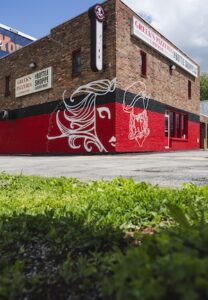 A low-angle shot shows the corner of a two-story brick building housing Greek's Pizzeria and The Bottle Shoppe. The lower half of the building is painted with a bold red stripe, featuring a large white line-art mural of a woman's face and a bearded man in a chef's hat. Signs for the pizzeria and liquor mart are visible on both sides of the building, and a patch of green grass is in the immediate foreground.