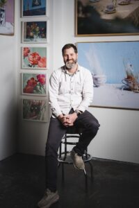 vertical portrait shows a man with a grey-flecked beard sitting on a metal barstool in an art gallery or studio. He wears a light-colored button-down shirt over a grey t-shirt, dark pants, and tan sneakers. He is smiling warmly with his hands clasped in his lap. Behind him, several framed pieces of art are displayed on white walls, including a large blue-toned print to his right and a vertical row of smaller colorful floral and abstract prints to his left.