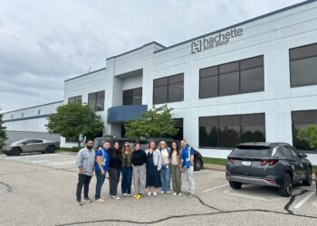 A group of ten people stand smiling together in a parking lot in front of a large, modern office building. The building has a sign that reads "Hachette Book Group." Two cars, including a silver Cybertruck and a dark SUV, are visible on either side of the group.
