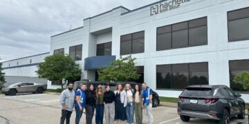 A group of ten people stand smiling together in a parking lot in front of a large, modern office building. The building has a sign that reads "Hachette Book Group." Two cars, including a silver Cybertruck and a dark SUV, are visible on either side of the group.