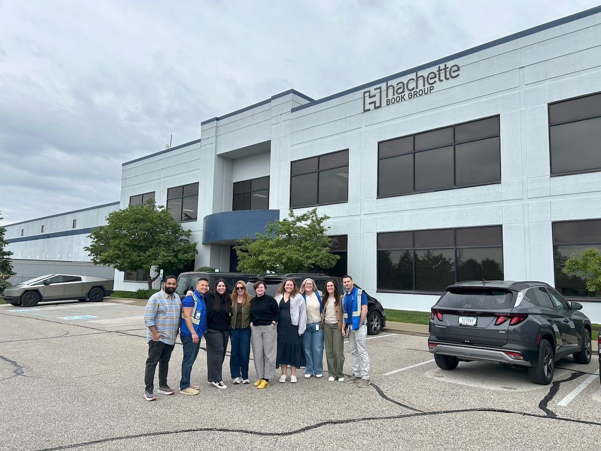 A group of ten people stand smiling together in a parking lot in front of a large, modern office building. The building has a sign that reads "Hachette Book Group." Two cars, including a silver Cybertruck and a dark SUV, are visible on either side of the group.