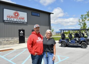 A man and a woman stand smiling in front of the Hurricane Golf Carts showroom. The man is wearing a red company hoodie and a baseball cap, and the woman is in a black top and blue jeans. Behind them is a grey industrial building with a large company sign and two blue street-legal golf carts parked in the lot.