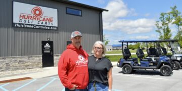A man and a woman stand smiling in front of the Hurricane Golf Carts showroom. The man is wearing a red company hoodie and a baseball cap, and the woman is in a black top and blue jeans. Behind them is a grey industrial building with a large company sign and two blue street-legal golf carts parked in the lot.