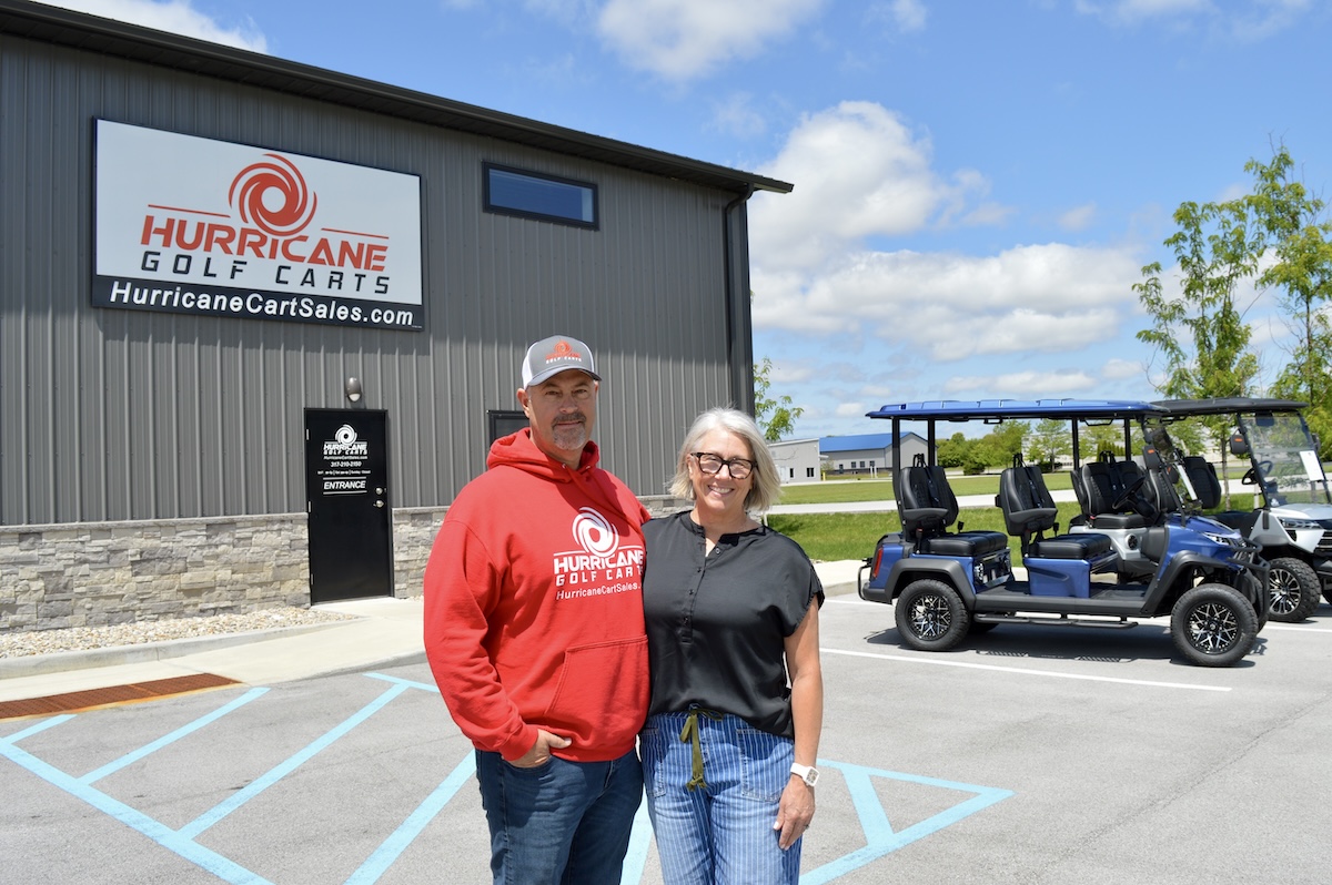 A man and a woman stand smiling in front of the Hurricane Golf Carts showroom. The man is wearing a red company hoodie and a baseball cap, and the woman is in a black top and blue jeans. Behind them is a grey industrial building with a large company sign and two blue street-legal golf carts parked in the lot.