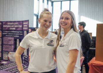 Two young women smiling and standing side-by-side at a presentation booth. They are both wearing white polo shirts with a "VITAFLEX SPORTS BANDAGES" logo. To their left is a purple display board titled "VITAFLEX: FUTURE OF WOUND CARE." The background shows a brightly lit indoor event space with large windows.