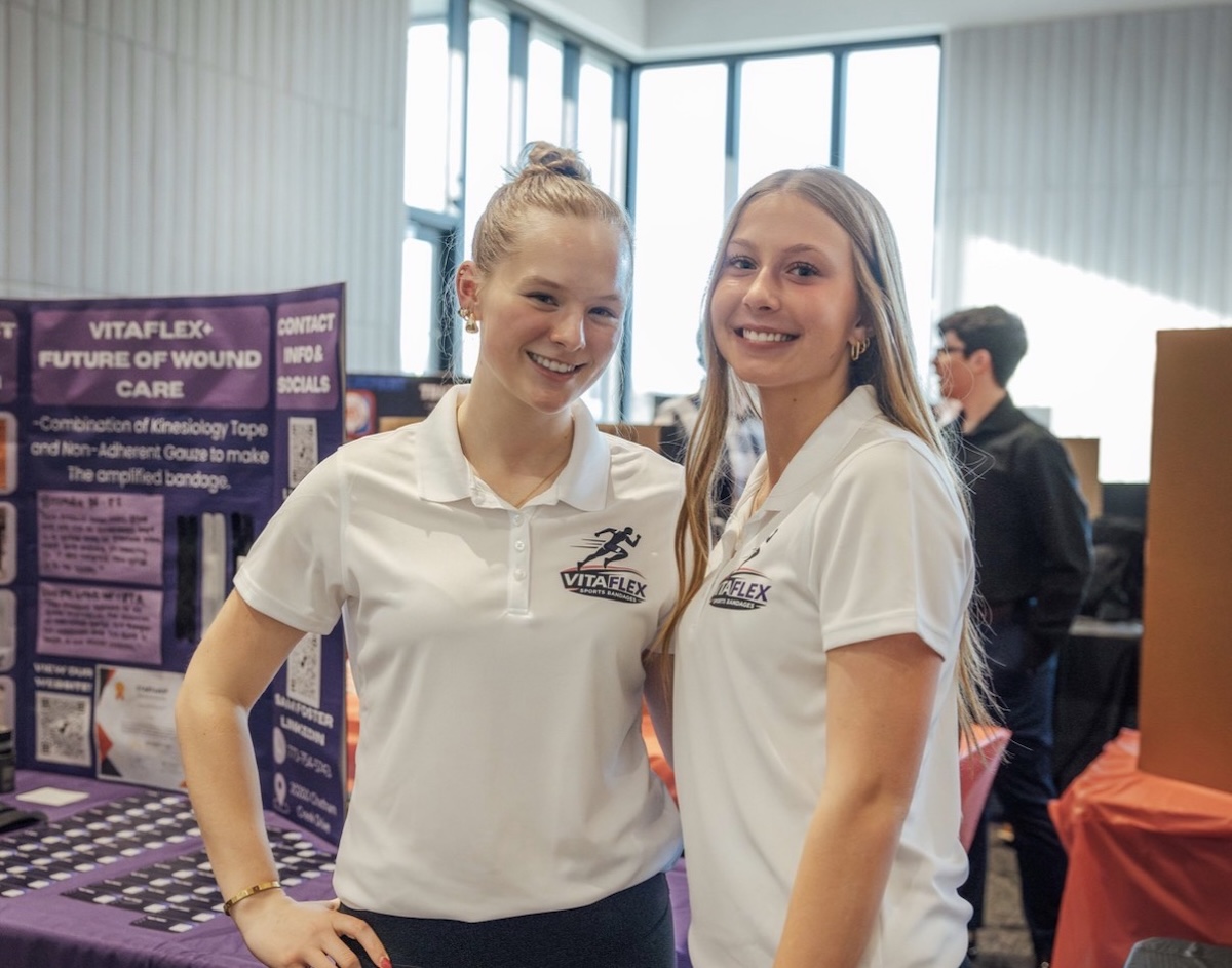 Two young women smiling and standing side-by-side at a presentation booth. They are both wearing white polo shirts with a "VITAFLEX SPORTS BANDAGES" logo. To their left is a purple display board titled "VITAFLEX: FUTURE OF WOUND CARE." The background shows a brightly lit indoor event space with large windows.