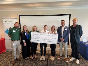 A group of seven people standing together in a conference room, holding a large ceremonial check for $2,000 from the Westfield Business Connection. The three women in the center hold the check, while four men stand on the ends. A "Westfield Chamber of Commerce" banner is visible in the background next to a large projector screen.