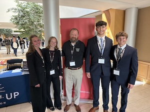 A group of five professionals posing for a photo in a bright, modern atrium. Two women on the left wear black suits, while a man in the center wears a black polo shirt and khaki pants. To the right, two young men wear dark suits and ties. All five are wearing conference lanyards. A red banner stands behind them, and a blue table with "UP" visible on the front is to the left.