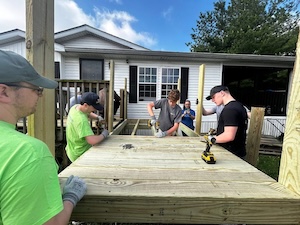 A close-up action shot of a volunteer group working on a wooden wheelchair ramp project. Three young men are actively using power drills to secure floorboards onto the ramp's frame. Two volunteers in the foreground wear bright lime green t-shirts and work gloves, while the house and a clear blue sky are visible in the background.