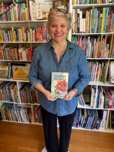 A woman with short blonde hair and glasses on her head stands smiling in front of floor-to-ceiling bookshelves. She is wearing a denim button-down shirt and black pants, and she is holding a book titled "Counterweights" by Jennifer Clement. The shelves behind her are packed with colorful children's books.