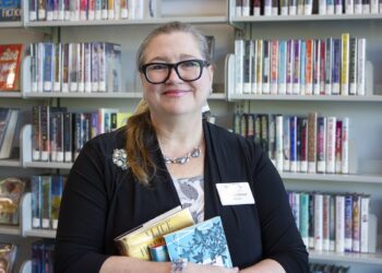 A woman with blonde hair, black-rimmed glasses, and a name tag that reads "Lintner" smiles while standing in front of library bookshelves. She is wearing a black cardigan over a floral top and is holding a small stack of books. The shelves behind her are filled with various fiction titles.