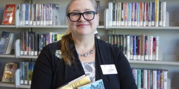 A woman with blonde hair, black-rimmed glasses, and a name tag that reads "Lintner" smiles while standing in front of library bookshelves. She is wearing a black cardigan over a floral top and is holding a small stack of books. The shelves behind her are filled with various fiction titles.
