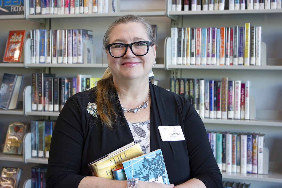 A woman with blonde hair, black-rimmed glasses, and a name tag that reads "Lintner" smiles while standing in front of library bookshelves. She is wearing a black cardigan over a floral top and is holding a small stack of books. The shelves behind her are filled with various fiction titles.