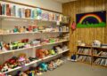 A brightly lit indoor play area features white shelves packed with a variety of toys, board games, and puzzles. To the right, two small wooden bookshelves are filled with children's books. A wood-paneled wall is decorated with a large green banner featuring a colorful rainbow. The carpeted floor is tidy, with several clear plastic bins holding smaller toy pieces.