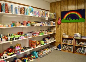 A brightly lit indoor play area features white shelves packed with a variety of toys, board games, and puzzles. To the right, two small wooden bookshelves are filled with children's books. A wood-paneled wall is decorated with a large green banner featuring a colorful rainbow. The carpeted floor is tidy, with several clear plastic bins holding smaller toy pieces.