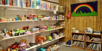 A brightly lit indoor play area features white shelves packed with a variety of toys, board games, and puzzles. To the right, two small wooden bookshelves are filled with children's books. A wood-paneled wall is decorated with a large green banner featuring a colorful rainbow. The carpeted floor is tidy, with several clear plastic bins holding smaller toy pieces.