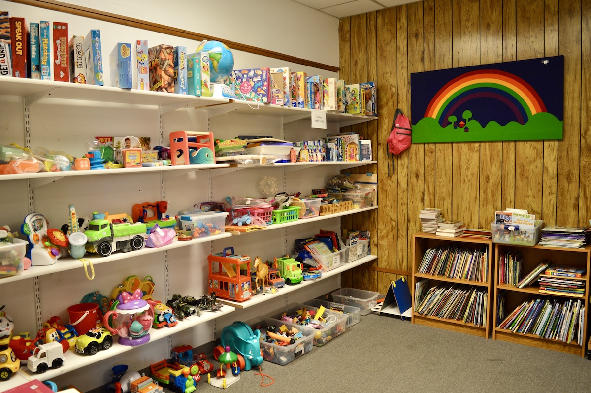 A brightly lit indoor play area features white shelves packed with a variety of toys, board games, and puzzles. To the right, two small wooden bookshelves are filled with children's books. A wood-paneled wall is decorated with a large green banner featuring a colorful rainbow. The carpeted floor is tidy, with several clear plastic bins holding smaller toy pieces.