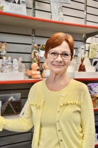 A woman with short red hair and glasses smiles warmly while standing in a gift shop. She is wearing a pale yellow sweater set with lace detailing. Behind her are white slatwall shelves filled with various decorative figurines, including angels, cherubs, and religious icons.
