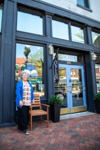 A full exterior shot of the "One More Chapter" bookshop facade featuring large black-painted wood panels, expansive clear windows, and double blue entry doors under a glass transom. A smiling woman with short silver hair, wearing a vibrant multi-colored crochet top and a bright blue cardigan, stands on a red brick sidewalk to the left of the entrance. Next to her is a wooden slatted armchair and a tall black planter with a green shrub. A black banner hanging over the door reads "OPEN" in bold white lettering.