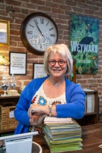 A medium shot of a smiling woman with short silver hair and red glasses, leaning her arms on a stack of books on a wooden checkout counter. She wears a blue cardigan over a colorful, patterned crochet top. Behind her, an exposed brick wall features a large round clock showing approximately 11:05, framed notices, and a promotional book poster for "Weyward" by Emilia Hart. A retail point-of-sale monitor is partially visible in the bottom left corner.
