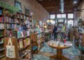 An interior shot looking down the narrow aisle of an indie bookstore with an exposed brick wall and high ceilings. Tall wooden bookshelves packed with books line the left wall under a green "NON-FICTION" sign. A canvas tote bag with text reading "Books and Coffee" hangs from a shelf foreground left. In the center, a round wooden table displays stacks of books on a teal and gold vintage patterned area rug. Several customers browse shelves and stand near a wooden checkout counter further down the aisle toward the bright storefront windows.