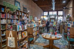 An interior shot looking down the narrow aisle of an indie bookstore with an exposed brick wall and high ceilings. Tall wooden bookshelves packed with books line the left wall under a green "NON-FICTION" sign. A canvas tote bag with text reading "Books and Coffee" hangs from a shelf foreground left. In the center, a round wooden table displays stacks of books on a teal and gold vintage patterned area rug. Several customers browse shelves and stand near a wooden checkout counter further down the aisle toward the bright storefront windows.