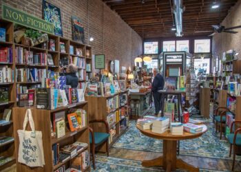 An interior shot looking down the narrow aisle of an indie bookstore with an exposed brick wall and high ceilings. Tall wooden bookshelves packed with books line the left wall under a green "NON-FICTION" sign. A canvas tote bag with text reading "Books and Coffee" hangs from a shelf foreground left. In the center, a round wooden table displays stacks of books on a teal and gold vintage patterned area rug. Several customers browse shelves and stand near a wooden checkout counter further down the aisle toward the bright storefront windows.