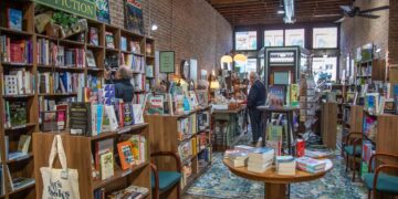 An interior shot looking down the narrow aisle of an indie bookstore with an exposed brick wall and high ceilings. Tall wooden bookshelves packed with books line the left wall under a green "NON-FICTION" sign. A canvas tote bag with text reading "Books and Coffee" hangs from a shelf foreground left. In the center, a round wooden table displays stacks of books on a teal and gold vintage patterned area rug. Several customers browse shelves and stand near a wooden checkout counter further down the aisle toward the bright storefront windows.