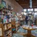 An interior shot looking down the narrow aisle of an indie bookstore with an exposed brick wall and high ceilings. Tall wooden bookshelves packed with books line the left wall under a green "NON-FICTION" sign. A canvas tote bag with text reading "Books and Coffee" hangs from a shelf foreground left. In the center, a round wooden table displays stacks of books on a teal and gold vintage patterned area rug. Several customers browse shelves and stand near a wooden checkout counter further down the aisle toward the bright storefront windows.