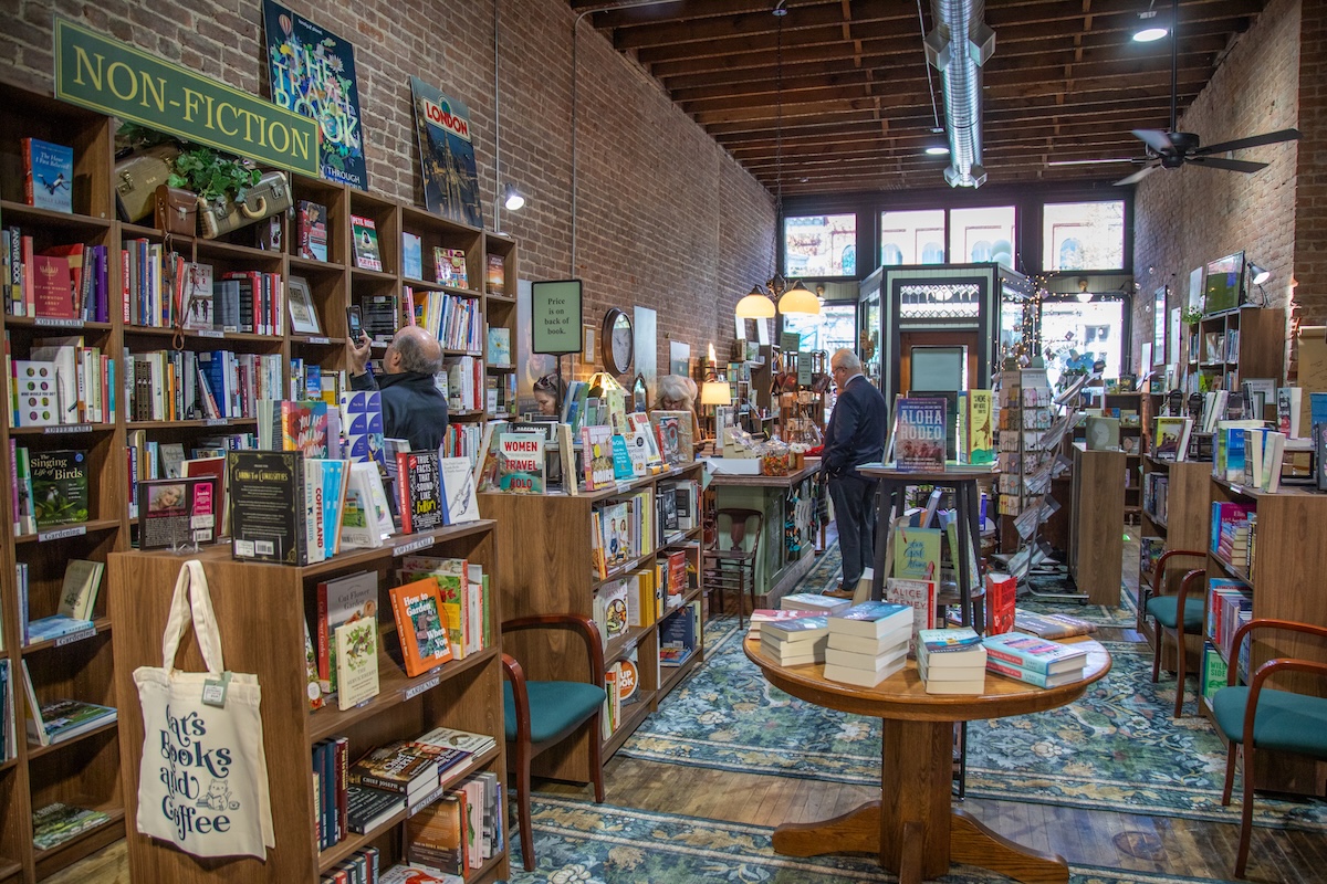 An interior shot looking down the narrow aisle of an indie bookstore with an exposed brick wall and high ceilings. Tall wooden bookshelves packed with books line the left wall under a green "NON-FICTION" sign. A canvas tote bag with text reading "Books and Coffee" hangs from a shelf foreground left. In the center, a round wooden table displays stacks of books on a teal and gold vintage patterned area rug. Several customers browse shelves and stand near a wooden checkout counter further down the aisle toward the bright storefront windows.