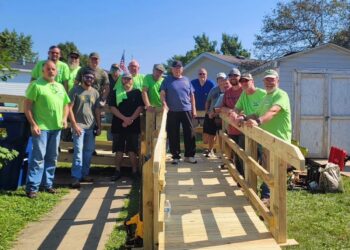 A large group of about 15 men stands on and around a newly constructed wooden wheelchair ramp outside a residential home. Several men are wearing bright lime green t-shirts. The group is smiling, with one man in a blue shirt standing at the top of the ramp. A small white shed and trees are visible in the background under a clear sky.
