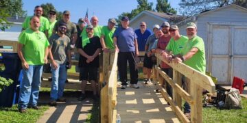 A large group of about 15 men stands on and around a newly constructed wooden wheelchair ramp outside a residential home. Several men are wearing bright lime green t-shirts. The group is smiling, with one man in a blue shirt standing at the top of the ramp. A small white shed and trees are visible in the background under a clear sky.