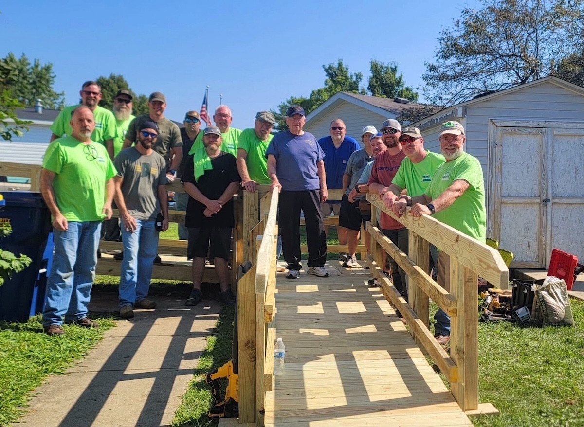 A large group of about 15 men stands on and around a newly constructed wooden wheelchair ramp outside a residential home. Several men are wearing bright lime green t-shirts. The group is smiling, with one man in a blue shirt standing at the top of the ramp. A small white shed and trees are visible in the background under a clear sky.