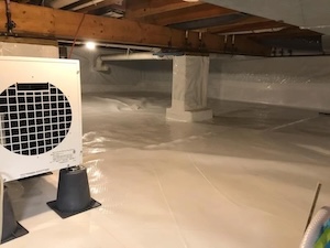 An interior shot shows a residential crawl space that has undergone complete moisture remediation and encapsulation. Bright white, heavy-duty plastic vapor barrier sheeting completely covers the dirt floor, foundation walls, and concrete support piers. On the left, a large white commercial crawl space dehumidifier stands elevated on black plastic blocks, showing a circular mesh vent. Exposed wooden floor joists and a large wooden support beam run across the low ceiling, illuminated by a single overhead light bulb.