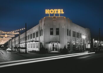A dramatic, long-exposure night photograph shows the Art Deco exterior of the Bottleworks Hotel. The building features white terra cotta walls and a glowing yellow neon "HOTEL" sign on the roof. To the left, strings of warm Edison bulbs hang over a brick street, creating a festive atmosphere. In the foreground, white light trails from a passing car zip across the dark asphalt, adding a sense of motion to the quiet city scene.