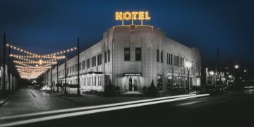 A dramatic, long-exposure night photograph shows the Art Deco exterior of the Bottleworks Hotel. The building features white terra cotta walls and a glowing yellow neon "HOTEL" sign on the roof. To the left, strings of warm Edison bulbs hang over a brick street, creating a festive atmosphere. In the foreground, white light trails from a passing car zip across the dark asphalt, adding a sense of motion to the quiet city scene.