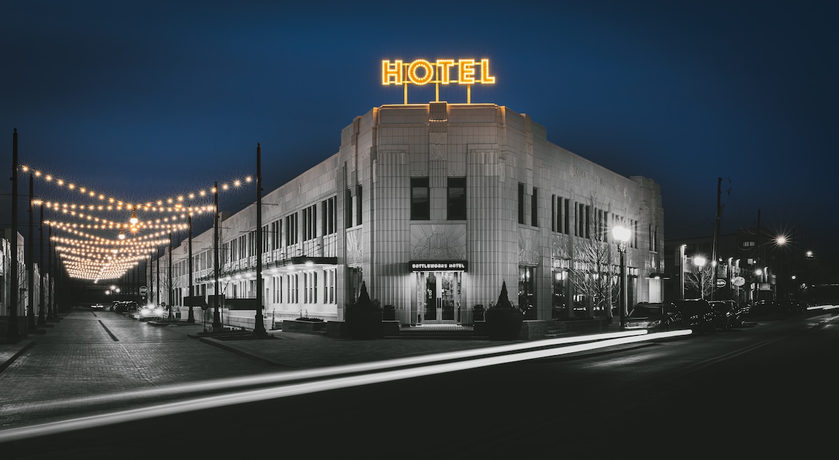 A dramatic, long-exposure night photograph shows the Art Deco exterior of the Bottleworks Hotel. The building features white terra cotta walls and a glowing yellow neon "HOTEL" sign on the roof. To the left, strings of warm Edison bulbs hang over a brick street, creating a festive atmosphere. In the foreground, white light trails from a passing car zip across the dark asphalt, adding a sense of motion to the quiet city scene.