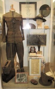 A glass museum display case showcases historical United States military artifacts from World War I. On the left, a full olive drab wool service uniform, including a high-collared tunic jacket and trousers, stands on a mannequin torso. The right shelves display various wartime artifacts, including a broad-brimmed campaign hat on a mannequin head, a trench art shell casing, framed photographs, documents, maps, and an informational roster placard.