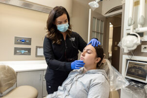 Clinical scene inside a modern treatment room at Fishers Dental Care. Dr. Shuttlesworth stands over a female patient who is resting back in a beige dental chair wearing a grey zip-up hoodie. The doctor wears a black uniform jacket, a light blue surgical face mask, and blue nitrile gloves. She is gently placing one hand on the patient's forehead and the other under her chin to assess her jaw and facial muscles. Specialized dental equipment, wall-mounted glove dispensers, and a computer monitor showing a dental scan are visible in the background.