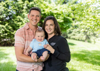 Outdoor three-quarter length portrait of a young family smiling together on a bright, sunny day with a lush green grass lawn and blurred trees in the background. The man on the left wears a coral and white striped short-sleeve button-down shirt and holds a baby boy. The baby wears a light blue striped t-shirt with fish graphics and navy blue shorts, smiling with his mouth open. The woman on the right has shoulder-length brown hair and wears a black zip-up athletic jacket featuring a white embroidered logo that reads "Fishers DENTAL CARE".