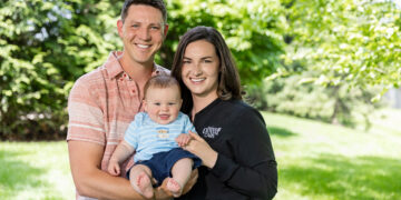 Outdoor three-quarter length portrait of a young family smiling together on a bright, sunny day with a lush green grass lawn and blurred trees in the background. The man on the left wears a coral and white striped short-sleeve button-down shirt and holds a baby boy. The baby wears a light blue striped t-shirt with fish graphics and navy blue shorts, smiling with his mouth open. The woman on the right has shoulder-length brown hair and wears a black zip-up athletic jacket featuring a white embroidered logo that reads "Fishers DENTAL CARE".