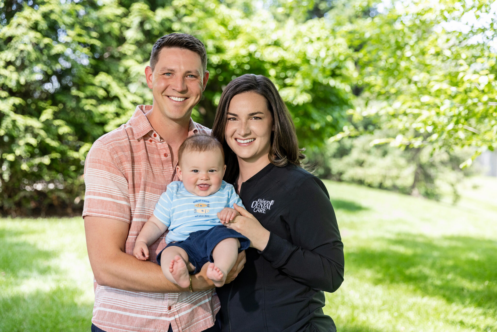 Outdoor three-quarter length portrait of a young family smiling together on a bright, sunny day with a lush green grass lawn and blurred trees in the background. The man on the left wears a coral and white striped short-sleeve button-down shirt and holds a baby boy. The baby wears a light blue striped t-shirt with fish graphics and navy blue shorts, smiling with his mouth open. The woman on the right has shoulder-length brown hair and wears a black zip-up athletic jacket featuring a white embroidered logo that reads "Fishers DENTAL CARE".