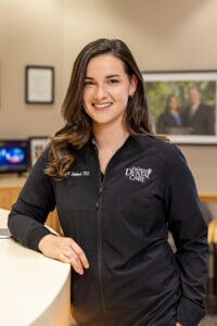 Three-quarter length indoor portrait of Dr. Shuttlesworth, DDS, leaning against a white curved reception desk. She has long, wavy brown hair draped over her shoulder and smiles warmly at the camera. She wears a black, zip-up uniform jacket with white embroidered cursive text reading "Dr. M. Shuttlesworth, DDS" on her right chest, and the block-letter logo for "Fishers DENTAL CARE" on her left chest. The blurred background shows a modern dental office lobby with framed wall art and a computer monitor.