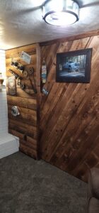 A room interior featuring accent walls made of rustic, stained wooden planks. The right wall has wood arranged in a diagonal pattern and holds a framed photo of a pickup truck. The left wall has horizontal planks decorated with car parts like a camshaft, timing chains, a turbocharger, a license plate, and a small decorative surfboard.