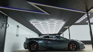 Wide side view of a dark grey McLaren 570S Spider sports car parked in a modern, professional detailing garage. The ceiling features a large, bright hexagon-shaped LED light grid centered above the vehicle, surrounded by dark slatted paneling and an accent lighting strip.