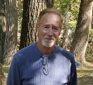 An outdoor portrait of a mature man with reddish-brown hair and a graying goatee, wearing a blue cable-knit sweater with glasses hooked into the collar, posing in front of a wooded forest background.