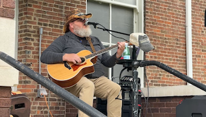 A musician with a full grey beard sits outside on a brick porch playing an acoustic guitar and singing into a microphone. He wears a grey long-sleeved shirt, tan pants, a patterned bucket hat, and sunglasses. A black metal railing runs along the foreground of the porch steps. A water bottle rests in a holder on his microphone stand, and a brick wall with a white-trimmed window serves as the background.