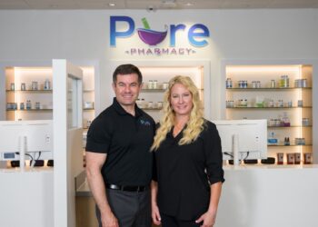 Indoor professional portrait of a man and woman standing together in a clean, modern pharmacy setting. The man on the left has short dark hair and wears a black polo shirt with a small "Pure" logo on the chest, paired with grey trousers. The woman on the right has long, wavy blonde hair and wears a black long-sleeve button-down blouse. They stand in front of a sleek white counter featuring desktop computer monitors. Behind them, glass product display shelves are recessed into white illuminated alcoves. A large backlit sign on the wall above features the company name, "Pure PHARMACY," with a colorful purple mortar and pestle integrated into the lettering.